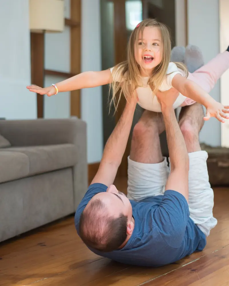 A father lying on a living room floor lifting his laughing daughter into the air