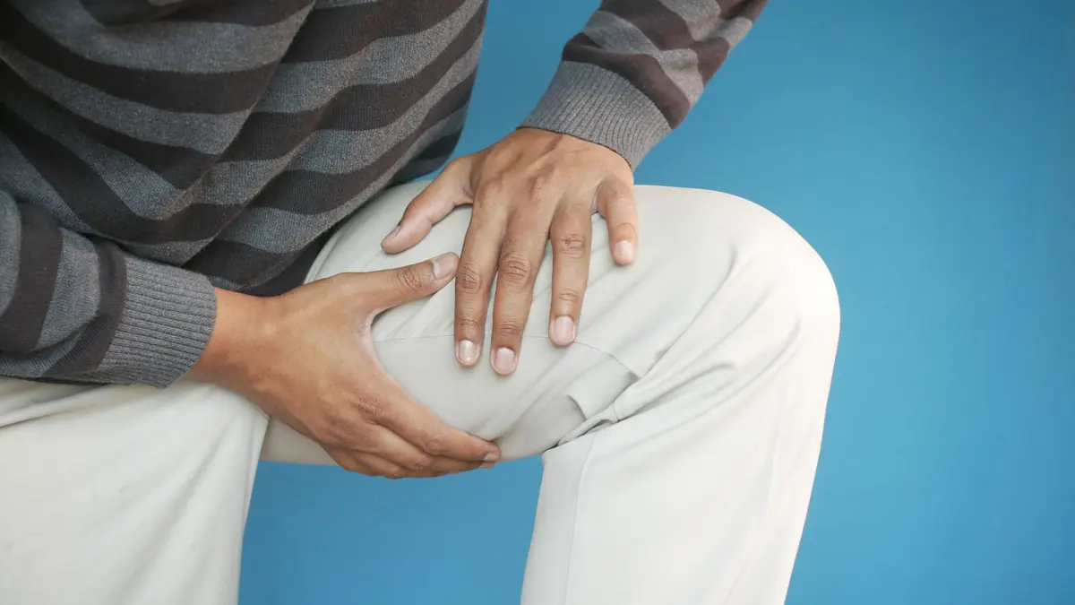 Man holding his thigh with both hands, standing against a blue wall