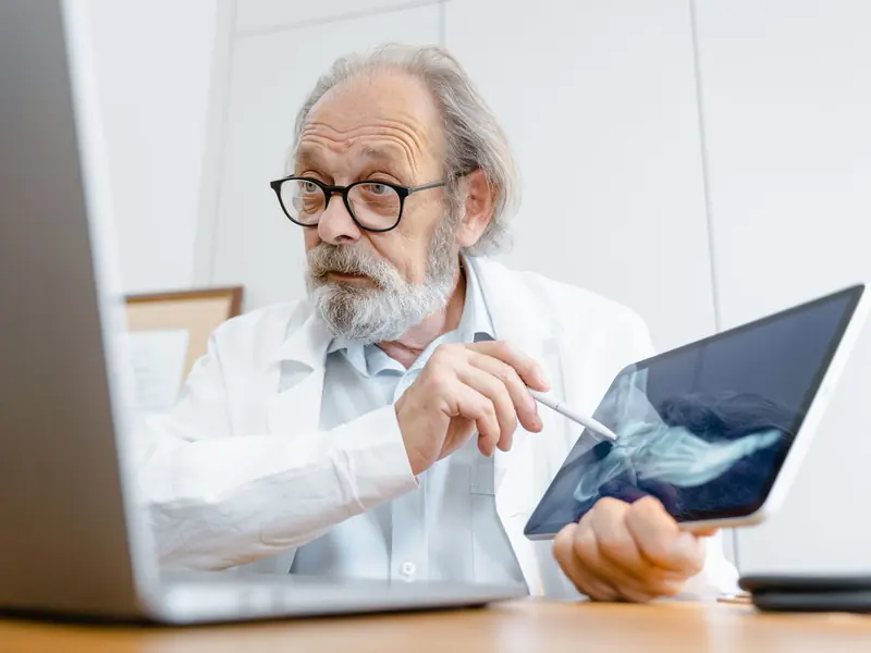 A doctor with white hair and glasses pointing to an X-ray image on a tablet